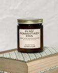 Amber glass jar candle labeled In My Romantasy Era on a stack of books with a white background.