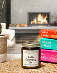 A jar candle with a white label that reads 'In My Romantasy Era', placed on a woven surface with books and a plant in the background.