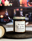 A jar candle with a label reading 'The Keeper of Keys and Grounds' next to a candle showing a smooth surface and wooden x wick on a wood surface with a plant and colored bottles in the background.