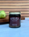 Candle jar labeled 'Last of the Harvest' by Gather the Flame on a blue surface with pears and a bottle of honey in the background.