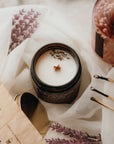 Candle in a black container with lavender flowers, surrounded by a book and matchsticks on a white surface.