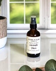 A brown amber spray bottle labeled 'Lavender Haze' by Gather In Flame Candle Co. on a white marble surface with a window and plant in the background.