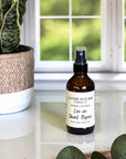 A brown amber spray bottle labeled 'Leo in Saint Tropez' by Gather In Flame Candle Co. on a white marble surface with a window and plant in the background.