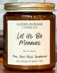Close up of a brown jar candle with a gold lid, labeled 'Let Us Be Menaces', placed on a wooden stand, with books and a window in the background.