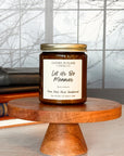 A brown jar candle with a gold lid, labeled 'Let Us Be Menaces', placed on a wooden stand, with books and a window in the background.