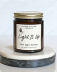 A jar candle with a gold lid, labeled 'Light It Up' by Gather In Flame Candle Co., with scents of peach, saffron, blackberry, and amber.
