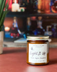 A jar candle with a gold lid, labeled 'Light It Up' by Gather In Flame Candle Co., with scents of peach, saffron, blackberry, and amber on an orange surface with bottles in the background.