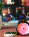 The surface of a candle with reddish color and gold glitter with a gold lid on an orange surface with a plant and bottles in the background.