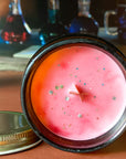 The surface of a candle with reddish color and gold glitter on an orange surface with bottles in the background.
