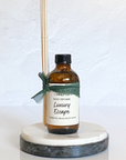 A reed diffuser with a brown bottle labeled 'Luxury Escape' on a round white marble platter, with a bamboo diffuser stick and green decorative ribbon.