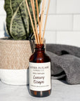 A brown glass reed diffuser bottle with a label reading "Luxury Escape", placed on a grey surface, surrounded by plant and towels with white tile background.