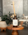 A brown glass reed diffuser bottle with a label reading "Luxury Escape", placed on a wood surface, with a plant and living room in the background.