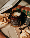 Candle in a jar labeled 'Midnight in the Library' on a table with books, roses, and glasses.