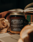 Candle jar labeled 'Midnight in the Library' by Gather of Flame on a surface with a rose and books.