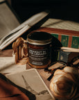 Candle labeled 'Midnight in theLibrary' on a surface with books and a rose.