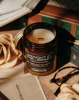 Jar of candle labeled 'Midnight in the Library' on a table with books and flowers