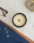 Topdown view of candle with wood wick on a textured surface with glasses, books and a clock in the background.