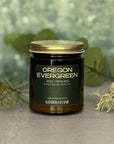Jar of 'Oregon Evergreen' candle with a gold lid on a grey surface with pine branches, moss and eucalyptus