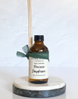A Reed diffuser with an amber glass bottle. There is a green ribbon and a label with the product name 'Prosecco Daydream' written on it placed on a marble stand.