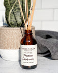 A brown glass reed diffuser bottle with a label reading "Prosecco Daydream", placed on a grey surface, surrounded by plant and towels with white tile background.