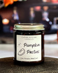 A jar candle with a label reading 'Pumpkin Pasties' on a wood surface with colored bottles in the background.