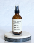 A brown bottle with a white label that reads 'GATHER IN FLAME CANDLE CO. ROOM & CAR SPRAY Relax & Release Pineapple Sage' on a white marble surface.