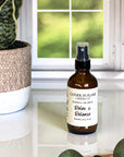 A brown amber spray bottle labeled 'Relax & Release' by Gather In Flame Candle Co. on a white marble surface with a window and plant in the background.