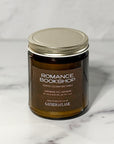 Close up of amber glass jar candle labeled Romance Bookshop with a white background.