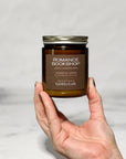 Amber glass jar candle labeled Romance Bookshop held in a hand with a white background.