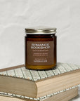 Amber glass jar candle labeled Romance Bookshop on top of a stack of books with a white background.