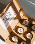 An open cardboard box labeled 'GATHER IN FLAME' with six small coconut wax candles arranged inside, each with a wooden wick.