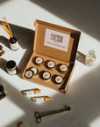 Scent discovery box surrounded by candles, reed diffusers, perfume oil rollers and match bottle on a white surface.