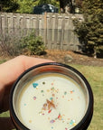 Hand holding a swiftie-inspired candle with colorful glitter sprinkles against a backyard background