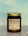 Candle jar labeled 'Snow at the Beach' with a gold lid on a blurred background