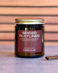 Candle jar labeled 'Spiced Plotlines' with a cinnamon stick on a wooden surface