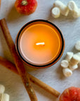 Overhead view of lit candle surrounded by cinnamon sticks, small pumpkins, and marshmallows on a light background