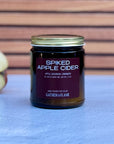 Jar of 'Spiked Apple Cider' candle with a purple label on a white surface with apple in the background