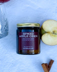 Flatlay of 'Spiked Apple Cider' candle with apples, a whiskey glass and cinnamon sticks on a white background