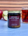 Jar of 'Spiked Apple Cider' candle with apples and a glass of cider and cinnamon stick in background.