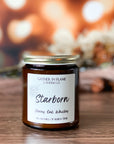A jar candle with a label and gold lid, bearing the name 'Starborn' and scent notes of Honey, Oak and Whiskey. The candle is placed on a wood surface with background of warm lights and flowers.