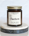 A jar candle with a dark label and gold lid, bearing the name 'Starborn' and scent notes of Honey, Oak and Whiskey. The candle is placed on a white marble surface.