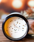 Candle surface with cotton wick and silver and gold glitter. The candle is placed on a wood surface with background of warm lights and flowers.