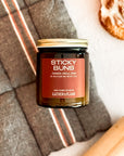 Candle labeled 'Sticky Buns' with a cinnamon roll and rolling pin on a light surface