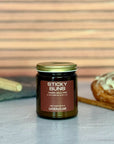 Jar of 'Sticky Buns' candle with a cinnamon roll, rolling pin and cinnamon stick on a wooden surface.