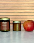 Two candles of different sizes labeled 'Sticky Buns' and 'Gather the Flame' next to an apple on a reflective surface with a wooden background.