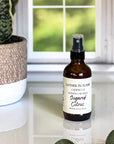 A brown spray bottle labeled 'GATHER IN FLAME CANDLE CO. ROOM & CAR SPRAY Sugared Citrus' placed on a white marble surface with a window and plant in the background.