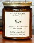 Close up of a jar candle labeled 'Tairn' by Gather in Flame Candle Co., placed on a wooden pedestal, with a window and books in the background.