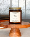 A jar candle labeled 'Tairn' by Gather in Flame Candle Co., placed on a wooden pedestal, with a window and books in the background.