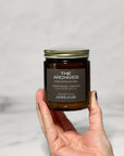 Amber glass candle Labeled The Archives held in a hand with a white background.