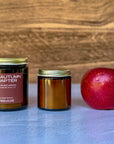 Two candles of different sizes labeled The Autumn Chapter with apple on wooden background
